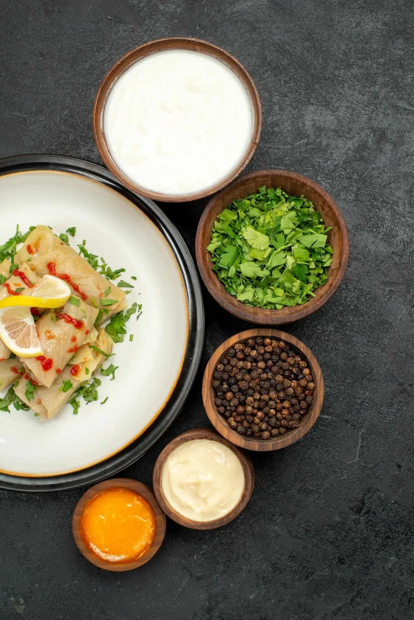 Draufsicht aus größerer Entfernung: Auf einem schwarzen Tisch stehen Schalen mit schwarzem Pfeffer, Sauerrahm, weißer und gelber Sauce sowie Kräutern sowie ein weißer Teller mit gefülltem Kohl mit Kräutern, Zitrone und Sauce.